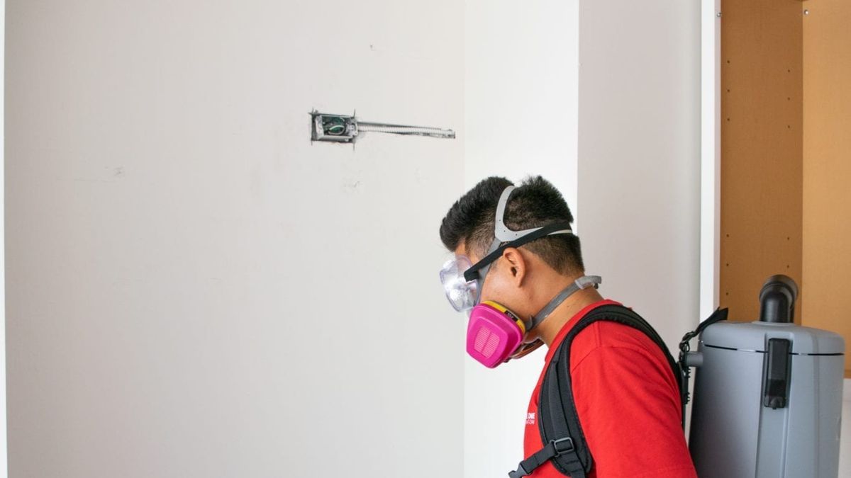 A Rescue One technician in a respirator HEPA-vacuuming a wall after water damage.