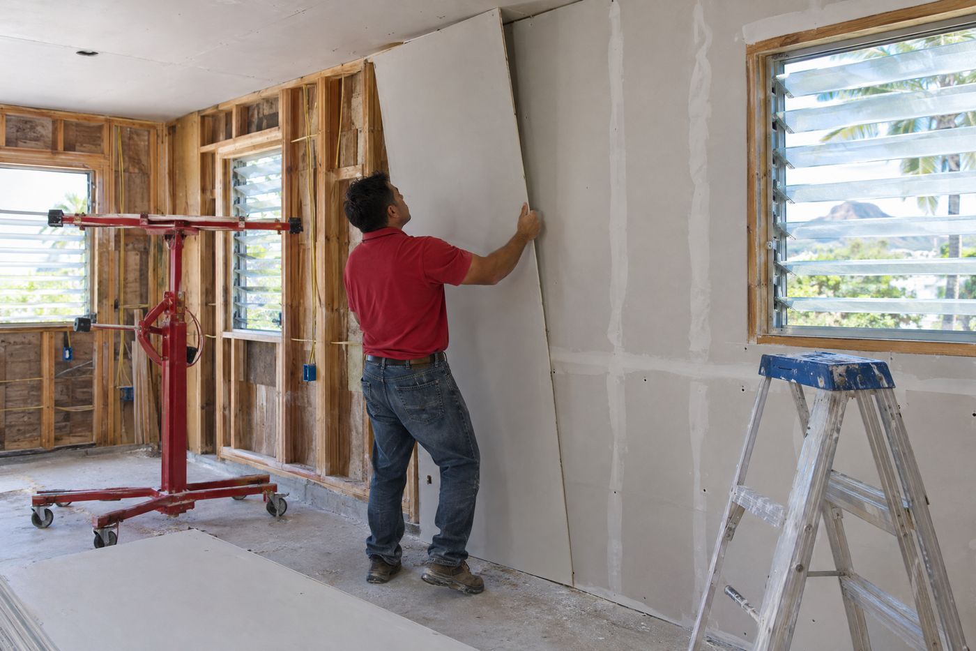 A worker lifting a new drywall sheet onto framing during a home rebuild in Oahu.