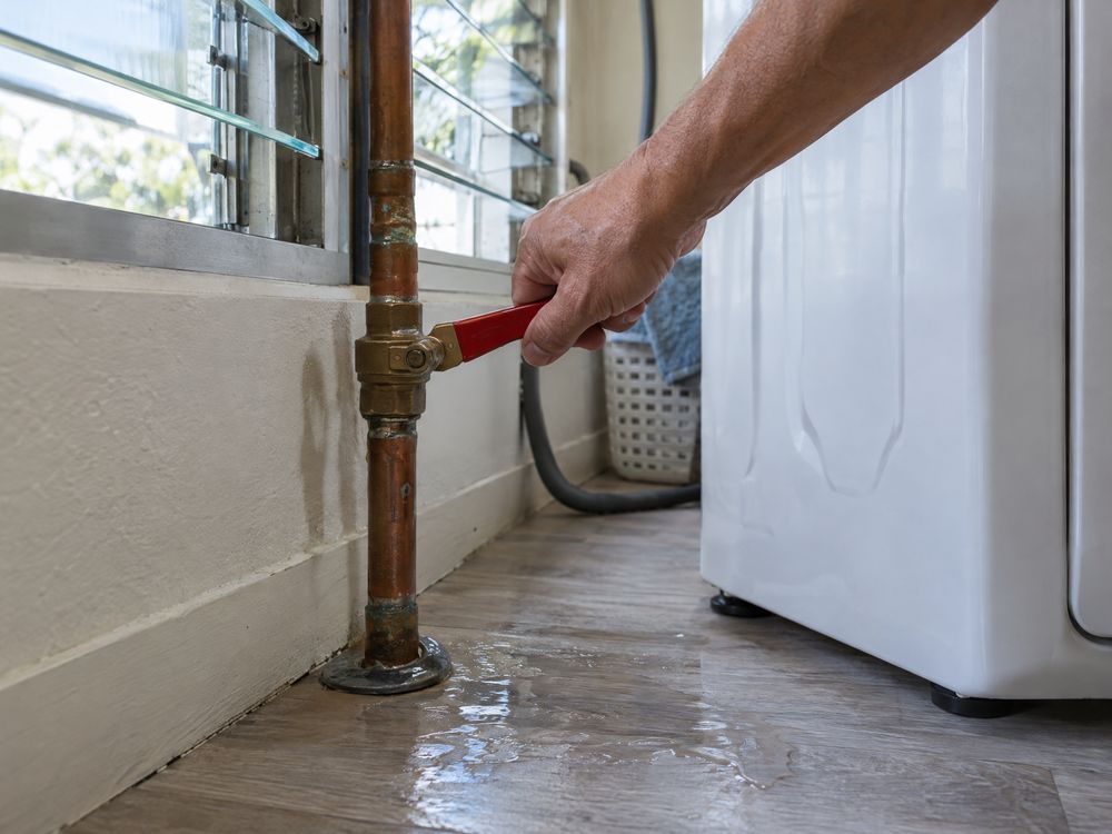 A homeowner turning off the main water shut-off valve beside a washing machine as water spreads across the floor.